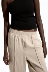 Pantalón de corte holgado color beige, confeccionado en sarga de mezcla de lyocell. Modelo de tiro alto con cintura fruncida, cierre decorativo y bolsillos laterales discretos. Piernas rectas con pliegues cosidos adelante y raya planchada atrás.