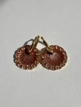 Aros colgantes con forma circular, hechos de resina color marrón con glitter y borde de strass. Cuentan con un arito de acero quirúrgico dorado.