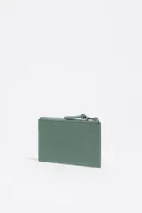 Monedero rectangular de Bimba y Lola, color verde esmeralda, con acabado brillante texturizado que simula piel de cocodrilo. Posee un cierre superior con cremallera y tirador de tela anudado. El logo metálico de la marca se encuentra centrado en la parte frontal.