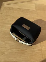 Neceser negro con cierre superior, forro interior y compartimento interno. Tiene un asa de mano con rayas horizontales blancas y negras.