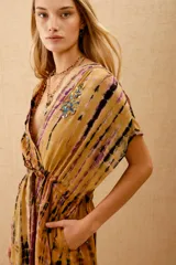 Vestido corto con estampado tie-dye en tonos mostaza, morado y negro. Presenta escote cruzado en V, mangas cortas tipo kimono y cintura ajustada con elástico. Tiene pequeños bordados de cuentas de colores en el escote.