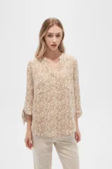 Blusa color beige con estampado de arabescos blancos, cuello mao, mangas 3/4 con lazos en los puños, cierre parcial con botones y bolsillos frontales.