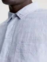 Camisa celeste de lino y algodón, de corte regular, manga larga, cuello en punta, cierre de botones oculto y bolsillo de parche en el pecho con monograma de Calvin Klein en la misma gama de tonos.