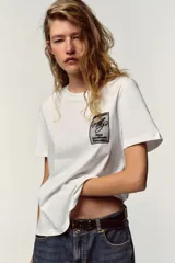 Camiseta blanca de algodón con cuello redondo y mangas cortas. Estampado frontal con texto "Meet me at Palais Lomtam, 7, Passage des Rêveries" enmarcado en un diseño ondulado.