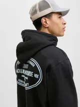 Buzo con capucha y manga larga, fit holgado, color negro. Confeccionado en tejido de punto circular loopback 100% algodón con interior cepillado. Presenta un estampado ovalado en la espalda con texto en blanco que incluye "ESTABLISHED IN COPENHAGEN 1990", "JACK&JONES WORLDWIDE MRK", "ORIGINALS CREATIVE DEPARTMENT DAILY BASICS" y "FROM VISION TO REALITY".