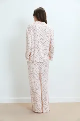 Pijama de dos piezas, compuesto por una camisa de manga larga y pantalón largo, ambos en tejido de viscosa color blanco con estampado integral de corazones rojos. La camisa tiene ribetes rojos en cuello, puños y abertura frontal. El pantalón tiene un fajín ancho color rojo liso.