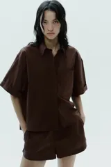 Camisa marrón de popelín con cuello solapa, manga corta con vuelta, bolsillo de plastrón en el pecho, bajo con aberturas laterales, pinzas en la espalda y cierre frontal con botones ocultos.