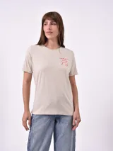Remera blanca de algodón con cuello redondo y manga corta. Presenta un pequeño bordado en el pecho izquierdo con una figura que parece ser una persona acostada en una tabla de surf o flotador.