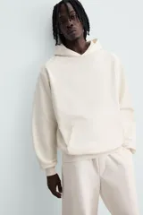 Sudadera con capucha color crudo, de corte relajado, confeccionada en tejido de algodón con interior rizado. Cuenta con cuello con capucha, manga larga, bolsillo frontal tipo canguro y acabados en rib.
