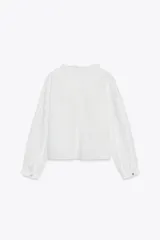Camisa blanca de cuello alto y manga larga, con detalle de entredós combinados a tono y cierre frontal con botones.
