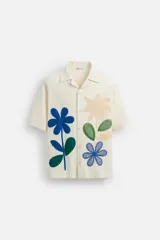 Camisa de manga corta color crudo, con cuello bowling, corte relaxed fit y cierre frontal con botones. Presenta apliques de parches con texturas combinadas a contraste en el delantero y aberturas laterales en el bajo.