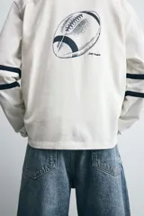Camiseta blanca de manga larga con cuello en pico. Tiene el número 12 bordado en el pecho y dos franjas horizontales en color azul marino en cada manga.