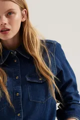 Camisa de denim azul con cuello clásico, cierre frontal con botones a presión, dos bolsillos con solapa abotonada en el pecho, mangas largas con puños abotonados y canesú en la espalda.