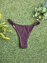 Bikini color marrón oscuro con corpiño tipo top texturizado, con hebilla ovalada marrón en el centro y tirantes finos. La bombacha es colaless entera con frunces en los costados. Incluye tazas soft removibles.