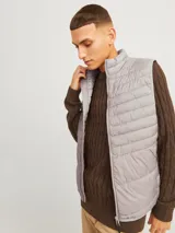Chaleco puffer gris con cuello alto y cierre frontal con cremallera. Tiene bolsillos laterales y diseño acolchado horizontal.