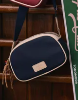 Bandolera de cuerina azul marino y beige con correa regulable azul y charm lateral decorativo. Tiene cierre superior y tag frontal con logo "The Brunch Club".