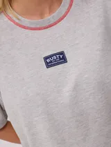 Remera gris de algodón con corte cropped y calce holgado. Tiene cuello redondo y mangas cortas con costuras reforzadas en color coral. Presenta un pequeño logo de la marca Rusty en el pecho.