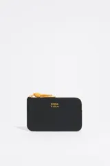 Monedero negro de cuero con cierre de cremallera y tirador de cordón amarillo. Incluye bolsillo interior y placa metálica con el logo de la marca.