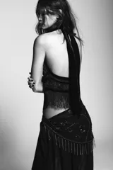 Top halter negro con cuello ajustable anudado, escote halter y hombros descubiertos. Presenta un panel central de terciopelo y apliques de abalorios brillantes en la parte delantera, con bajo asimétrico terminado en flecos largos.