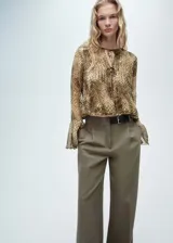 Blusa de poliéster con estampado de serpiente en tonos marrones, beige y negro. Tiene mangas largas acampanadas, cuello redondo con cierre de lazo en la parte delantera y diseño recto.