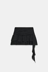 Falda pantalón negra de tiro bajo con cintura drapeada y bajo acabado en volantes superpuestos. Presenta un lazo largo y fino que cae desde el lateral.