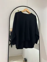Sweater negro de punto con cuello redondo, puños y bajo elásticos. El frente es más corto que la parte trasera y tiene botones en la espalda.