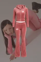 Conjunto deportivo de terciopelo rosa compuesto por un buzo con capucha y cierre frontal, y un pantalón acampanado. Ambos con logo estampado en blanco.