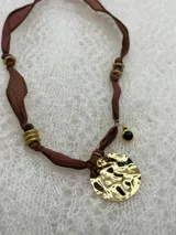 Collar con cinta de seda color marrón, adornado con cuentas doradas y un dije circular dorado con textura irregular.