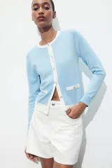 Chaqueta celeste de punto con cuello redondo y manga larga. Tiene cierre frontal con botones y detalle de falsos bolsillos delanteros con ribete blanco.