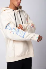 Buzo canguro amarillo ocre oversize de algodón rústico con capucha, cordones negros con letras blancas, bolsillo frontal y puños y cintura acanalados. Presenta estampados en el frente y las mangas.