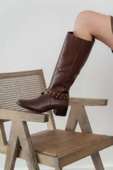 Bota de cuero color marrón con caña alta, taco cuadrado y punta fina. Presenta una correa con ojales metálicos dorados alrededor del tobillo.