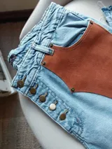 Pantalón de jean celeste con cintura elastizada, corte recto, bolsillos laterales y aplique de cuero marrón en el frente. Presenta botones metálicos en los laterales y etiqueta de cuero en la parte posterior.