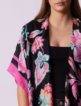 Kimono tejido con estampado floral multicolor, terminación de ruedo con borlas y mangas cortas con borde celeste.