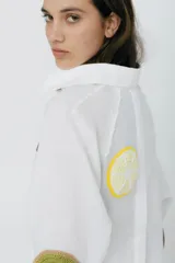 Camisa blanca de lino con corte raglán, detalles desflecados y apliques de frutas tejidas a crochet. Abertura trasera con botones.