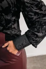 Camisa negra de terciopelo con motivos florales, cuello mao, mangas abullonadas, detalles plisados en el frente, botones forrados y bordes con puntilla.