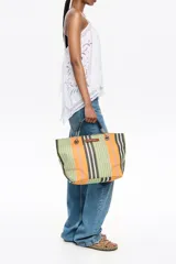 Bolso tote de tela a rayas verticales en tonos verde oliva, lila, blanco y rosa. Tiene doble asa de mano de cuero marrón y logo de la marca en etiqueta de cuero.