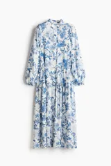 Vestido largo blanco con estampado floral en tonos azules. Tiene cuello mao, botones ocultos en la parte delantera, mangas largas abullonadas con puños elásticos, cinturón fino extraíble y costura fruncida debajo de la cadera para dar vuelo.
