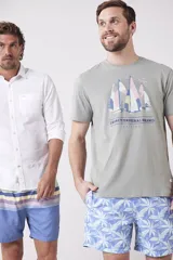 Remera de manga corta color verde grisáceo con cuello redondo. Presenta un estampado frontal de veleros sobre un fondo circular con líneas verticales, y texto bordado debajo.