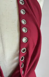 Vestido corto ajustado color rojo oscuro, estilo halter con escote pronunciado en V que se ajusta con ojales metálicos a lo largo del borde. El ruedo también presenta ojales metálicos.