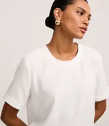 Blusa blanca de poliéster con cuello redondo, mangas cortas, cierre trasero en forma de gota, pequeñas aberturas laterales y acabados en pespuntes en las mangas y el cuello.