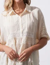 Blusa color crema de crepe con cuello a la base con solapa, escote en V con abertura central y botones. Delantera con detalles plisados y apliques en crochet. Manga corta con terminación fruncida y ruedo en broderie.