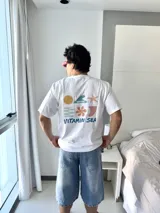 Remera blanca de manga corta con cuello redondo y corte holgado. Presenta un estampado gráfico en la espalda con elementos marinos (sol, ola, estrella de mar, flor, etc.) y el texto "VITAMIN SEA" y "BROKERS".