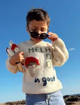 Sweater tejido a mano en lana merino color crema, con cuello redondo y mangas largas. Presenta un aplique bordado de un helado de dos bochas y texto bordado con la frase "Help is good".