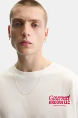 Camiseta blanca de corte relajado, con cuello redondo y mangas cortas. Presenta estampados combinados en contraste en la parte delantera y trasera con la frase "Gourmet Groove LLC".