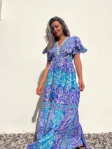Vestido largo de seda con estampado paisley en tonos turquesa, azul y blanco. Tiene escote en V, mangas cortas con volados y talle imperio con cordón ajustable.