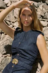 Vestido midi de denim azul oscuro, con cuello redondo y sin mangas. Presenta detalle de frunces en la cintura, cinturón elástico con hebilla dorada y cierre lateral con cremallera oculta. Tiene falsos botones delanteros.