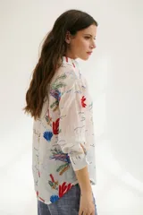 Camisa beige de manga larga con cuello clásico y cierre frontal con botones. Presenta bordados con motivos florales y geométricos en tonos azules, rojos y verdes.