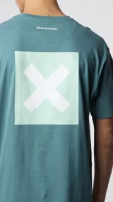 Camiseta celeste de algodón orgánico con cuello redondo y manga corta. Estampado en la espalda con un cuadrado turquesa y una cruz blanca en el centro.