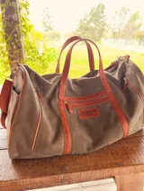 Bolso de viaje verde de cuero vacuno, con detalles en cuero marrón. Tiene doble asa de mano y correa desmontable para el hombro, cierre superior con cremallera, bolsillos exteriores e interiores.