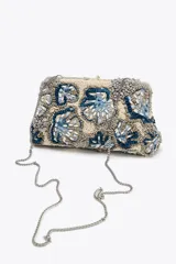 Bolso de mano tipo clutch con cuerpo cubierto de abalorios en tonos beige, azul y gris. Tiene un diseño floral y un cierre imantado. Incluye correa metálica extraíble y bolsillo interior con cremallera.